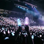 A sea of glowing K-pop lightsticks held aloft in a dark indoor arena, viewed from a low angle looking upward toward the stage lights.