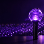 A hero product shot of a spherical globe-shaped K-pop concert lightstick — clear domed top over a matte-black cylindrical handle with a subtle ribbed grip — glowing with a soft purple-violet light from within. The lightstick stands slightly off-center on a dark reflective surface. Behind it, a dreamy bokeh sea of thousands of matching purple lights fills a darkened concert arena, softly out of focus. Cinematic studio lighting with a gentle purple rim light and a cool highlight on the glass dome. Premium, editorial, high-detail, photorealistic. Moody dark background with rich purple and deep-navy tones, a touch of warm glow. Clean negative space in the upper-left third for a title overlay. 16:9, high resolution.