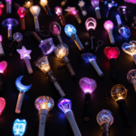 A dark, moody flat-lay of dozens of colorful glowing official K-pop lightsticks in different shapes — hearts, stars, moons, orbs and crystal-cut designs — representing the wide variety of lightsticks covered in the database below.